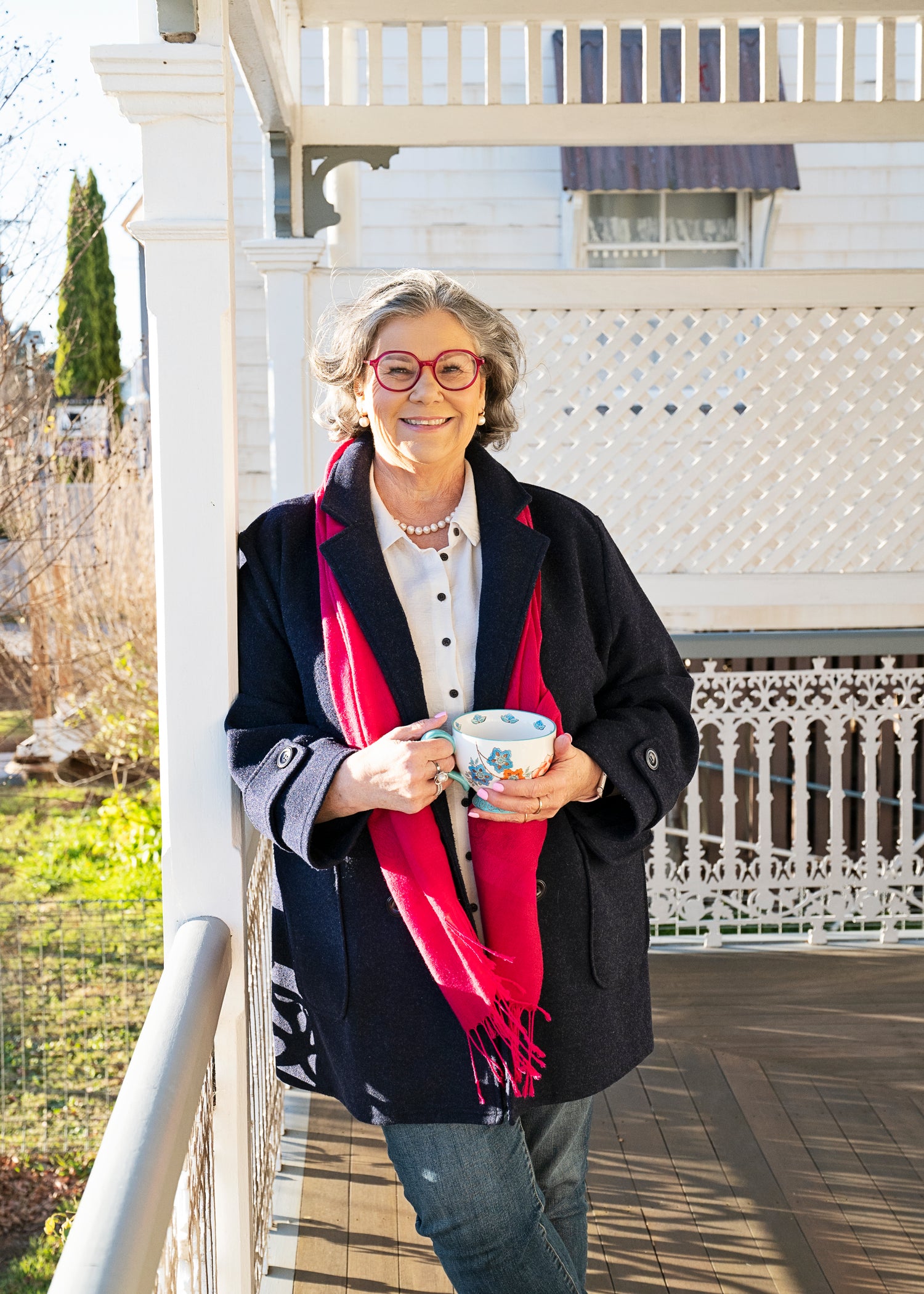 Author Susan Mackie holding a cup on a porch with a white railing and house in the background